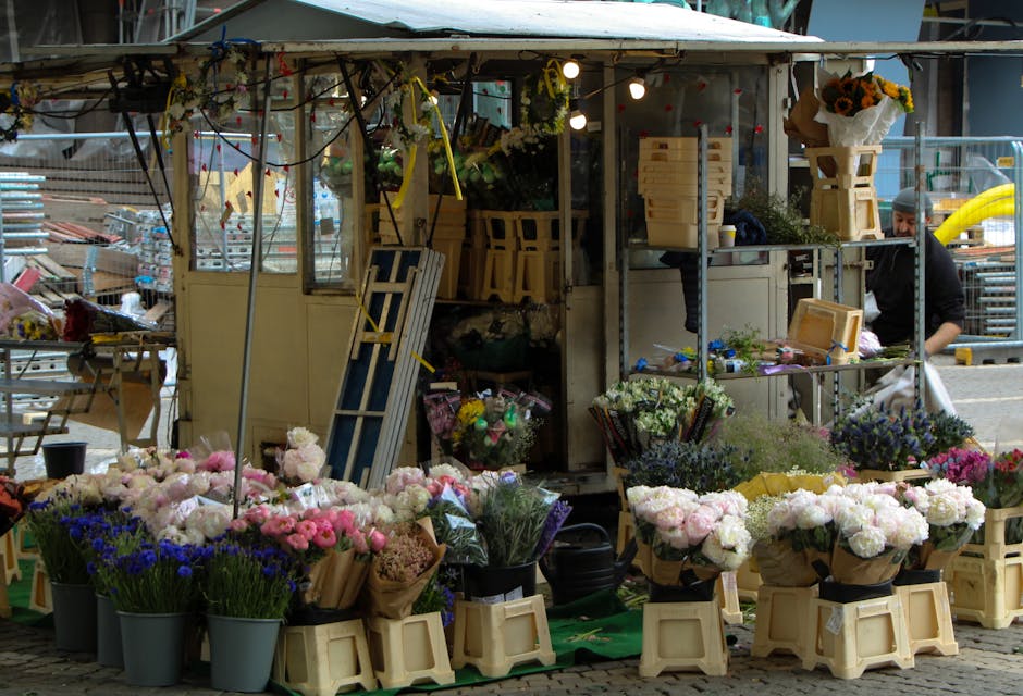A vibrant outdoor flower stall featuring an assortment of fresh flowers arranged in black buckets and wrapped bouquets with pink, white, purple, and blue blooms. The display includes peonies, hydrange