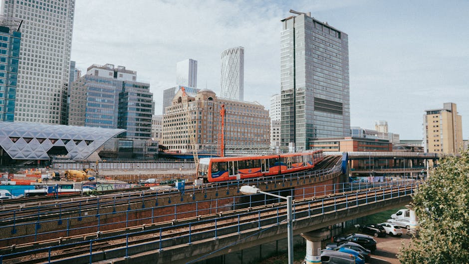 A vibrant orange and red tram moving along elevated railway tracks in a modern urban area with tall glass skyscrapers and contemporary architecture. The tram is centered in the image, with its sleek d