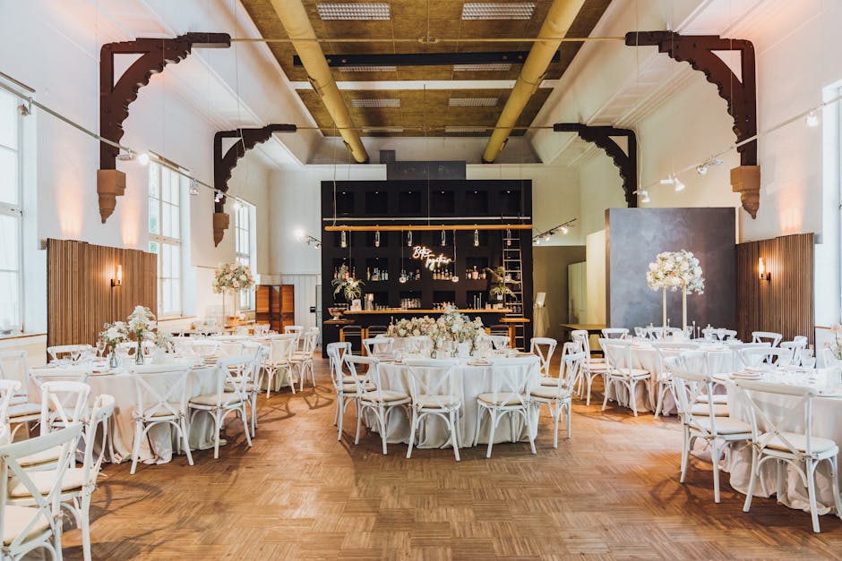 A spacious event hall set up for a wedding reception, featuring round tables draped with white tablecloths arranged in a semicircular pattern. Each table is decorated with elegant floral centerpieces 