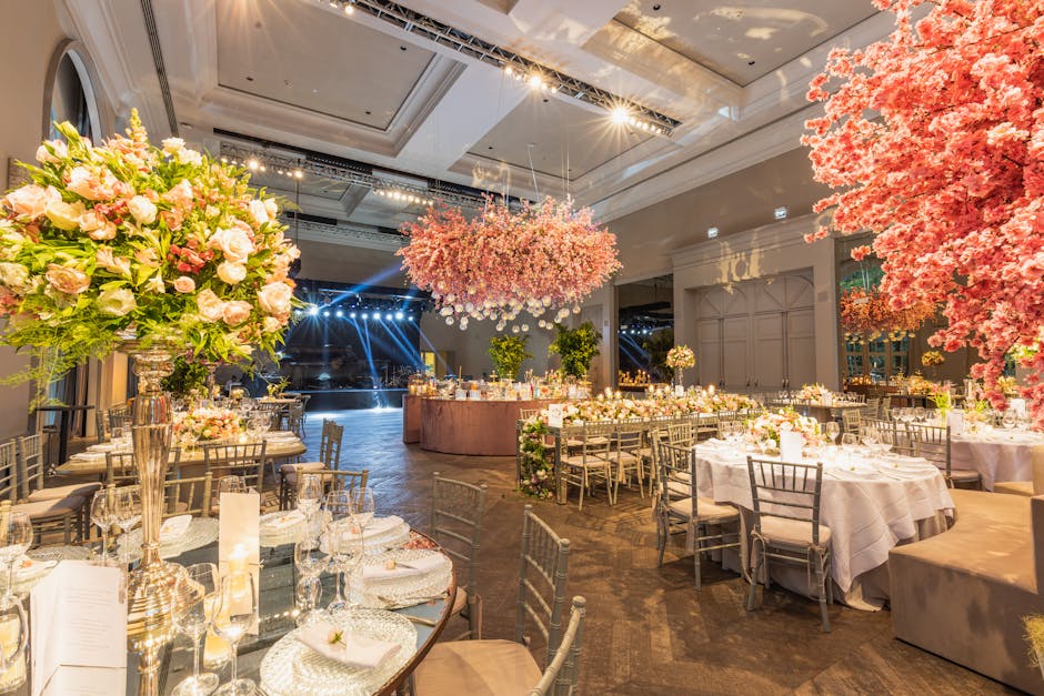 An elegantly decorated wedding reception venue with a high ceiling and spacious layout, featuring multiple round tables draped in white tablecloths and set with fine glassware, cutlery, and floral cen