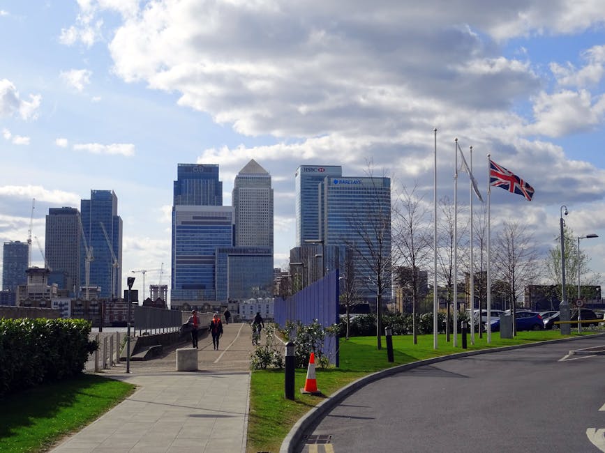 A view of Canary Wharf's modern skyline featuring tall office buildings with glass facades, set against a partly cloudy sky. In the foreground, a landscaped area with a paved pedestrian walkway leads 
