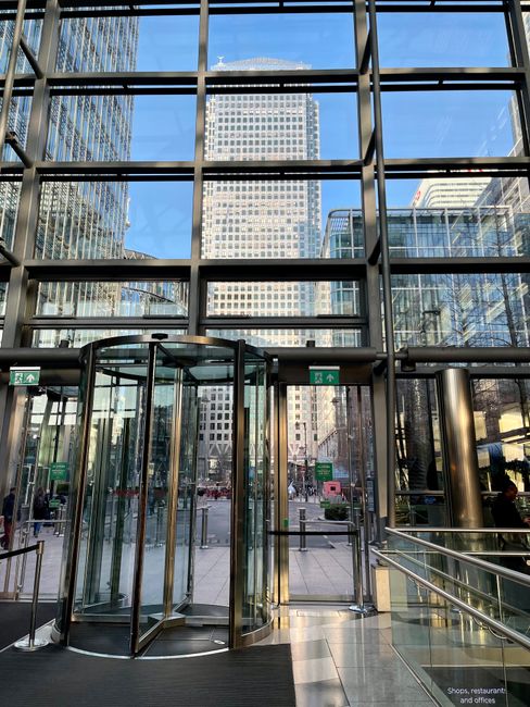 A large, modern glass entrance with a revolving door situated within a high-rise building featuring expansive floor-to-ceiling windows. Bright daylight filters through the clear glass, revealing a bus