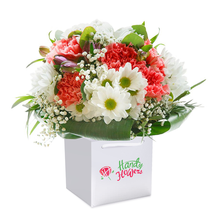 A stunning, abundant bouquet is thoughtfully arranged in a modern white square gift bag, photographed against a flawless white backdrop-a composition crafted with true Blackwall flair. The heart of the bouquet features volleys of soft coral and vivid red carnations, their plush, ruffled petals layered richly for depth and movement. Crisp white daisies with refreshing green-yellow centers are nestled among the carnations, shining with brightness and warmth reminiscent of sunlit mornings along the Poplar High Street. Tracing lines of pure, star-like baby's breath are woven throughout the arrangement, their gentle clusters lightening the palette and adding airy complexity. Around the base, smooth, deep green foliage and bold, broad leaves wrap like a collar, providing elegant support and framing the flowers as if in a living embrace. On the left, a shy cluster of unopened buds tinged with purple hints at promise and new beginnings, mirroring the ever-changing riverside scene of Blackwall. The white carrier bag, designed for both style and function, is topped with sturdy handles, perfect for gifting or doorstep delivery. With no distraction from logos or text, the focus is on the natural beauty and vivid artistry that define Blackwall Florist's work. Every stem, colour, and texture is carefully chosen, creating a gift that radiates freshness, joy, and the feeling of home in this lively East London community-ideal for birthdays, anniversaries, or simply brightening someone's day with a taste of Blackwall's floral charm.
